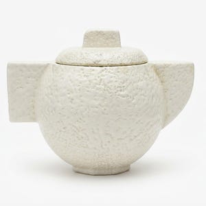 Op de afbeelding: Een ronde, gebroken witte theepot met een getextureerd, kraterachtig oppervlak. Het heeft een bijpassend deksel met een kleine, vierkante knop, een breed, plat handvat aan de linkerkant en een korte, hoekige tuit aan de rechterkant op een voet.