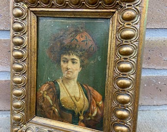 May include: A framed portrait of a woman wearing a red and green hat with a gold trim. The portrait is set in a gold frame with a beaded design.