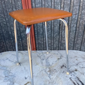 May include: A vintage, square-topped stool with a brown seat and chrome legs. The stool has a classic design, with a padded seat and four slender, curved legs. The legs are made of shiny chrome and have small, white rubber feet.