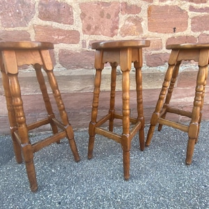 May include: Three wooden bar stools in a light brown finish. Each stool has a round seat, turned legs, and a footrest. The stools are arranged in a row against a brick wall.