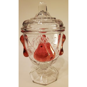 May include: A clear glass sugar bowl with a lid and pear-shaped handles. The bowl is decorated with an etched design of pears and leaves. The handles are a deep red color.