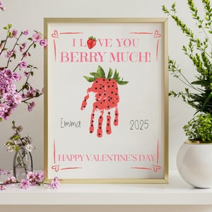 May include: A framed print with a gold border features a handprint transformed into a strawberry. The text reads "I Love You Berry Much!" and "Happy Valentine's Day." The name "Emma" and the year "2025" are also included.