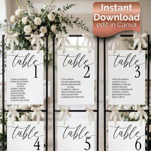 May include: A set of six white table number signs with black text, each adorned with a cream-colored bow. The signs are suspended from a black metal frame, with guest names listed below the table numbers. The top right corner has the text "Instant Download edit in Canva".