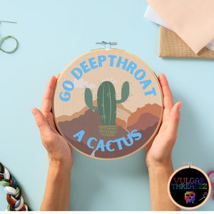 May include: A round embroidery hoop with a brown background and a green cactus in a pot. The text "Go Deep Throat A Cactus" is written in blue on the hoop.