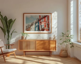 May include: A mid-century modern living room with a wooden cabinet, a white armchair, and a framed print of colorful row houses. The room is decorated with houseplants and has a large window with natural light.