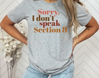 Op de afbeelding: Een grijs T-shirt met de tekst "Sorry, ik spreek geen sectie 8" in bruine en oranje letters.