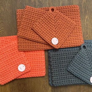May include: Three sets of crocheted pot holders. One set is orange, one is coral, and one is gray. Each set has two pot holders and a small tag with the word "Love" on it.