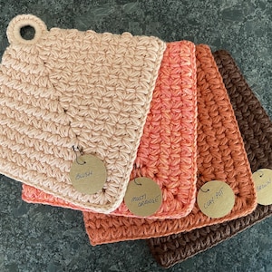 May include: Four crocheted pot holders in different shades of orange and brown. The pot holders are labeled with the colors "Blush", "Multi Orange", "Clay Pot", and "Brown".