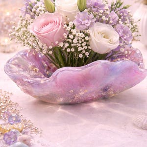 May include: A floral arrangement featuring pink and white roses, lavender flowers, and baby's breath in a decorative, iridescent, shell-shaped bowl. The bowl has a pink and purple color scheme with gold accents. The arrangement is set on a light pink surface.