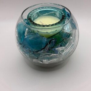 Puede incluir: Un portavelas de cristal transparente con borde turquesa y una vela amarilla en su interior. El soporte est&aacute; lleno de trozos de vidrio azules y verdes. El portavelas es redondo y se apoya sobre una superficie blanca.