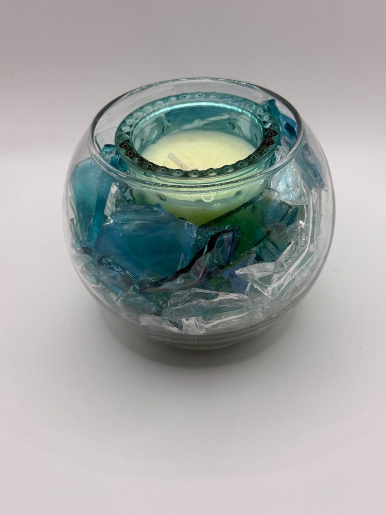 Puede incluir: Un portavelas de cristal transparente con borde turquesa y una vela amarilla en su interior. El soporte est&aacute; lleno de trozos de vidrio azules y verdes. El portavelas es redondo y se apoya sobre una superficie blanca.