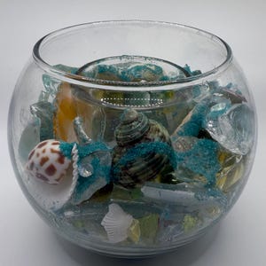 May include: A clear glass bowl filled with seashells, sea glass, and turquoise glitter. The bowl has a round shape and is filled with various sizes and colors of shells and glass. The overall theme is coastal decor.