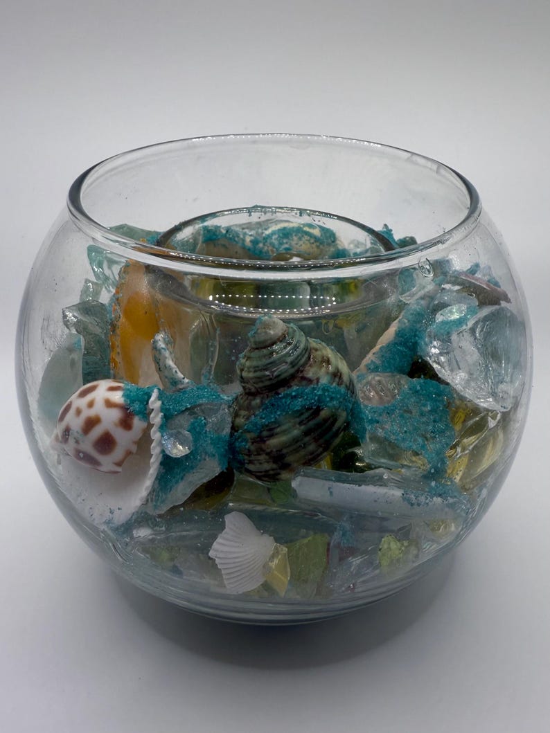 May include: A clear glass bowl filled with seashells, sea glass, and turquoise glitter. The bowl has a round shape and is filled with various sizes and colors of shells and glass. The overall theme is coastal decor.