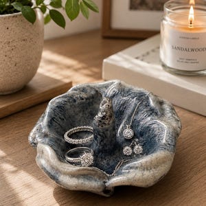 Può includere: Un portagioie in ceramica blu e bianca con una superficie strutturata. Il piatto contiene un anello, una collana e orecchini. Una candela accesa e una pianta in vaso sono sullo sfondo.