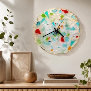 May include: A circular wall clock with a textured, translucent white resin base embedded with colorful shards of red, green, blue, and yellow glass. The clock features a thin gold-toned scalloped edge and three black hands at the center.
