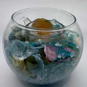 May include: A clear glass bowl filled with decorative elements. Inside, there's a smaller glass container holding a yellow object. The bowl is filled with blue-green sea glass, seashells, and other decorative items, creating a coastal theme.