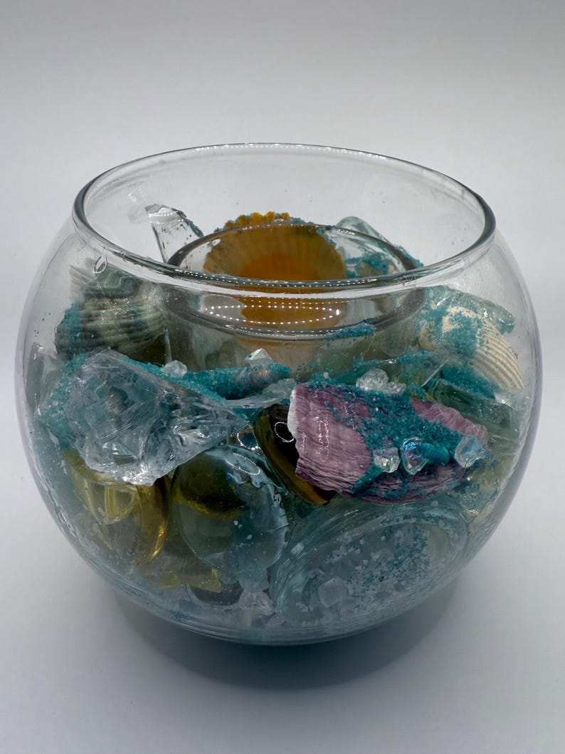 May include: A clear glass bowl filled with decorative elements. Inside, there's a smaller glass container holding a yellow object. The bowl is filled with blue-green sea glass, seashells, and other decorative items, creating a coastal theme.