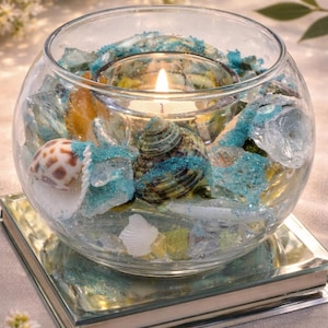 May include: A clear glass bowl filled with seashells, blue sand, and colorful glass pieces, with a lit candle in the center. The bowl sits on a mirrored surface, creating a reflective effect. A decorative item for home decor.