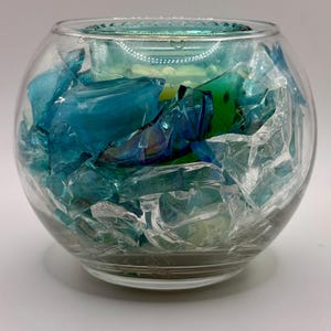 Puede incluir: Un cuenco de vidrio transparente lleno de fragmentos de vidrio azules y transparentes. El cuenco tiene forma redondeada y una superficie ligeramente texturizada. Los fragmentos de vidrio son de varios tonos de azul y transparente.