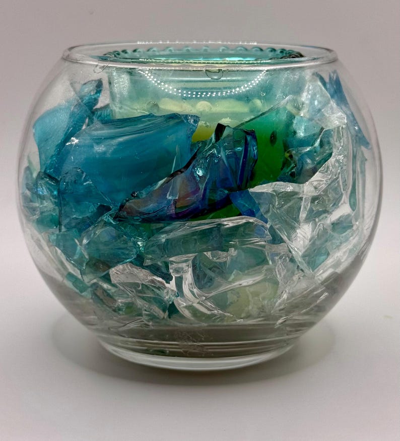 Puede incluir: Un cuenco de vidrio transparente lleno de fragmentos de vidrio azules y transparentes. El cuenco tiene forma redondeada y una superficie ligeramente texturizada. Los fragmentos de vidrio son de varios tonos de azul y transparente.