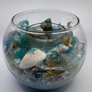 May include: A clear glass bowl filled with seashells, sea glass, and blue sand. The bowl has a round shape and is filled with various colors of sea glass, including blue, purple, and clear. Several seashells are visible.