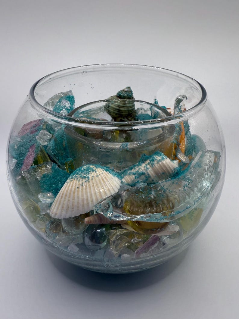 May include: A clear glass bowl filled with seashells, sea glass, and blue sand. The bowl has a round shape and is filled with various colors of sea glass, including blue, purple, and clear. Several seashells are visible.