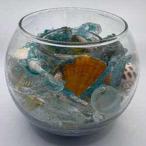 May include: A clear glass bowl filled with seashells, sea glass, and blue glitter. The bowl contains a large orange scallop shell, several white and brown seashells, and a clear marble. The contents are arranged on a bed of sand.