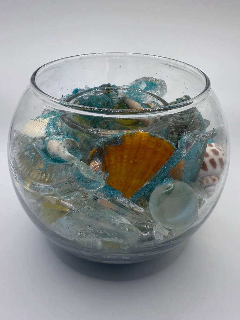 May include: A clear glass bowl filled with seashells, sea glass, and blue glitter. The bowl contains a large orange scallop shell, several white and brown seashells, and a clear marble. The contents are arranged on a bed of sand.