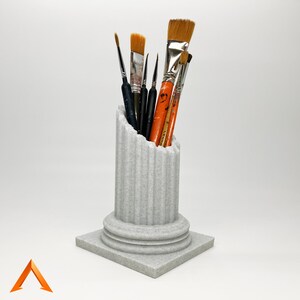 May include: A white ceramic column shaped like a Greek pillar with a fluted design. The column is filled with a variety of paint brushes, including some with orange handles.