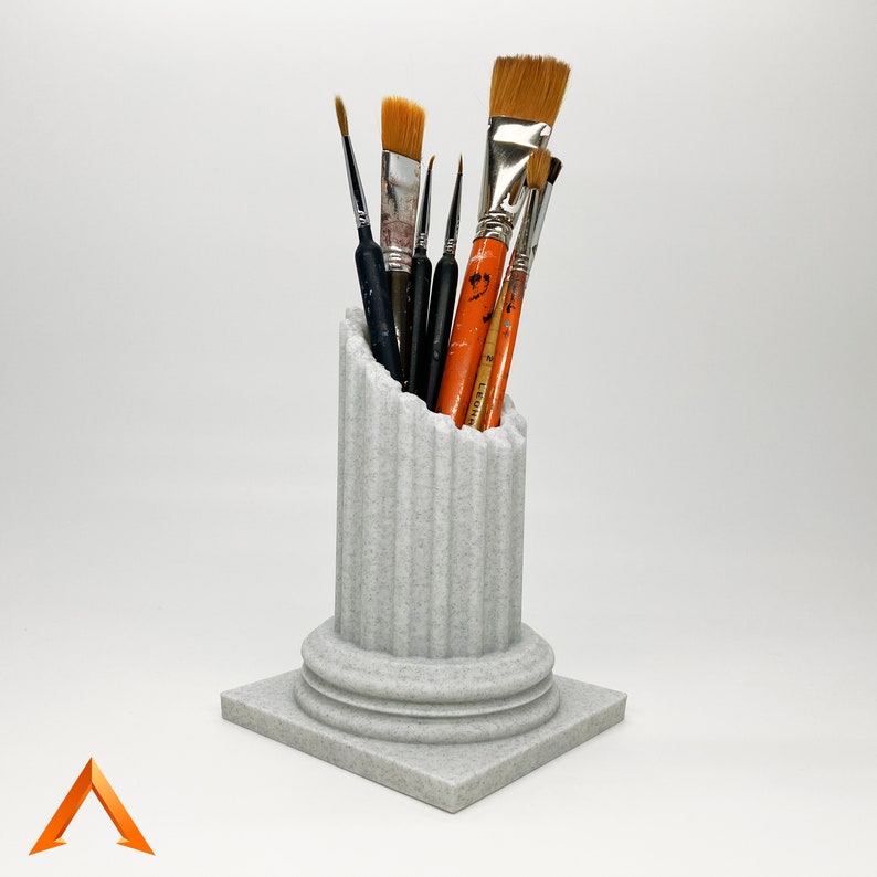 May include: A white ceramic column shaped like a Greek pillar with a fluted design. The column is filled with a variety of paint brushes, including some with orange handles.