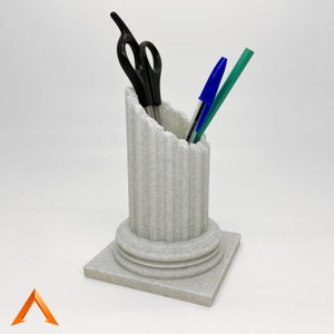 May include: A white ceramic desk organiser shaped like a column with a fluted design. It holds a pair of black scissors and two blue and green pens.