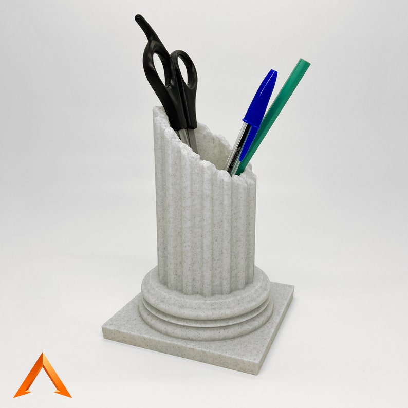 May include: A white ceramic desk organiser shaped like a column with a fluted design. It holds a pair of black scissors and two blue and green pens.