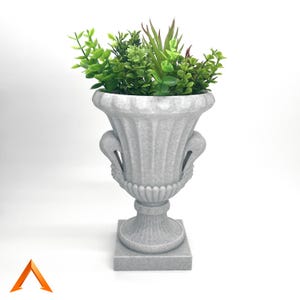 Op de afbeelding: Een witte keramische urnenplanter met een vierkante basis en sierlijke details. De urn is gevuld met verschillende groene planten.