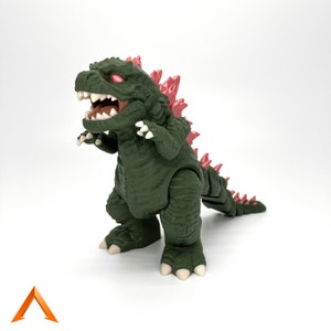 Op de afbeelding: Groene en rode plastic Godzilla-figuur met een wijd open mond en scherpe tanden. De figuur heeft een stekelige rug en een lange staart.