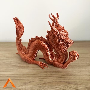 May include: A copper-coloured dragon figurine with intricate details. The dragon is curled up with its head turned to the side and its claws holding a small orb. The figurine is made of a smooth, metallic material.
