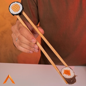Peut inclure: Une paire de baguettes en bois tenant un rouleau de sushi avec du saumon et de l'avocat. Les baguettes ont un anneau en forme de sushi à une extrémité.