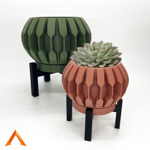 May include: Two geometric patterned plant pots with black metal stands. The larger pot is green and the smaller pot is orange. The smaller pot has a succulent plant inside.