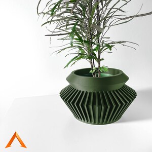 Peut inclure: Un pot de plante vert foncé avec un motif géométrique et une plante verte feuillue. Le pot a un design unique en couches avec un large dessus ouvert et une base avec des lignes verticales inclinées. La plante a de longues feuilles fines.