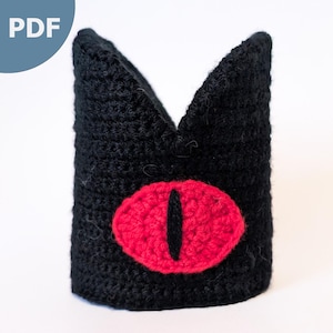 May include: A black crocheted crown with a large, red eye design. The eye is oval-shaped with a black vertical pupil. The crown has two pointed peaks and is made of a textured yarn. The PDF text is in the upper left corner.