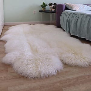 Puede incluir: Una gran alfombra de piel de oveja blanquecina con una textura peluda se encuentra sobre un suelo de madera. La alfombra tiene una forma irregular y cubre una parte importante del espacio del suelo. La alfombra está cerca de una cama con cabecero morado y una mesita.