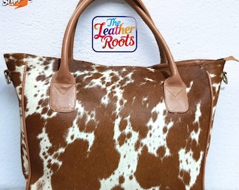May include: A brown and white cowhide tote bag with brown leather handles and trim. The bag has a zip closure and a rectangular shape. The text "The Leather Roots" is visible on the bag.