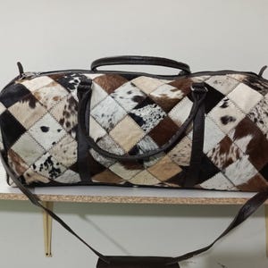 May include: A large duffel bag with a patchwork design. The bag features a diamond pattern of black, white, brown, and tan cowhide. It has a dark brown leather handle and a detachable shoulder strap. The bag is designed for travel.