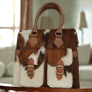 May include: A brown and white cowhide handbag with two front pockets and a top handle. The bag features brown leather accents, including the handle, pocket flaps, and straps. Gold-colored buckles secure the pockets.
