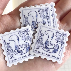Peut inclure: Quatre autocollants en papier blanc avec des illustrations bleues et blanches de serpents. Chaque autocollant a un bord festonné et dit "Année du Serpent" en texte bleu.