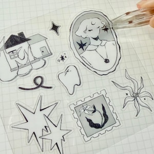 Könnte beinhalten: Eine Sammlung klarer Aufkleber mit verschiedenen Schwarz-Weiß-Illustrationen. Designs umfassen ein Haus, ein Porträt, einen Zahn, Sternausbrüche und eine Blume. Eine Hand verwendet eine Pinzette, um einen Aufkleber vom Bogen zu ziehen.