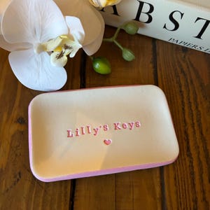 Peut inclure: Un plateau vide-poche rectangulaire blanc cassé aux coins arrondis avec un bord peint en rose. Le centre du plateau présente le texte rose en relief "Lilly's Keys" suivi d'un petit symbole de cœur rose centré sous le texte.