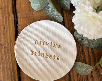 May include: A round, off-white trinket dish with a textured, diagonal ribbed surface. The center features the gold-colored embossed text "Olivia's Trinkets" in a clean, sans-serif font. The dish has a smooth, raised edge and a flat base.
