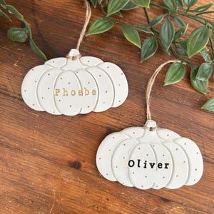 May include: Two white ceramic pumpkin ornaments with brown polka dots and jute twine hangers. One pumpkin features the name Phoebe in gold lettering, and the other features the name Oliver in black lettering, both with a smooth, matte finish.