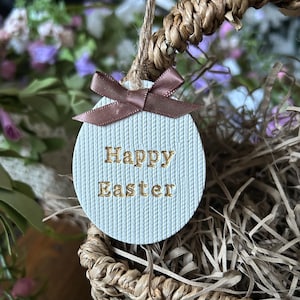 May include: A circular, pale blue Easter decoration with the words "Happy Easter" in gold lettering. It has a brown ribbon and hangs from a woven basket. The background includes greenery and flowers, creating a festive scene.