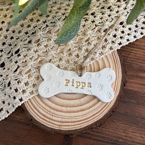 May include: A white bone-shaped ornament featuring embossed paw prints across the surface and the name Pippa printed in gold lettering. The ornament includes a natural jute twine loop for hanging, providing a simple and functional design.